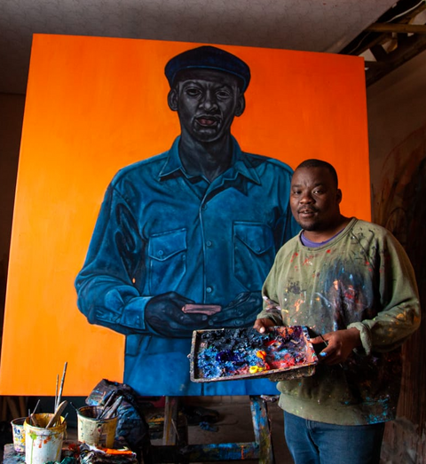 Ein Foto von einem unserer Künstler. Sein Name ist Khaya Sineyile und auf dem Foto kann man ihn neben einer seiner Kunstwerke sehen.