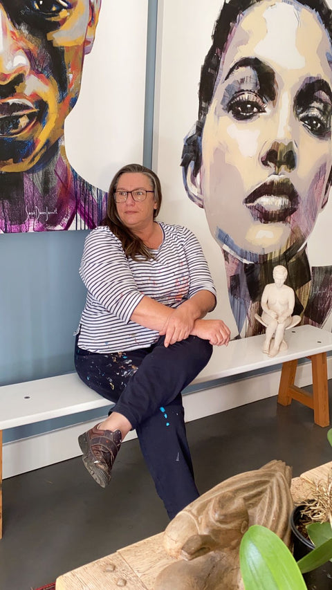 Das Bild zeigt die südafrikanische Künstlering Sarah Danes Jarrett wie sie vor 2 ihrer Kunstwerke aus ihrer Kunstreihe "Women Series" sitzt